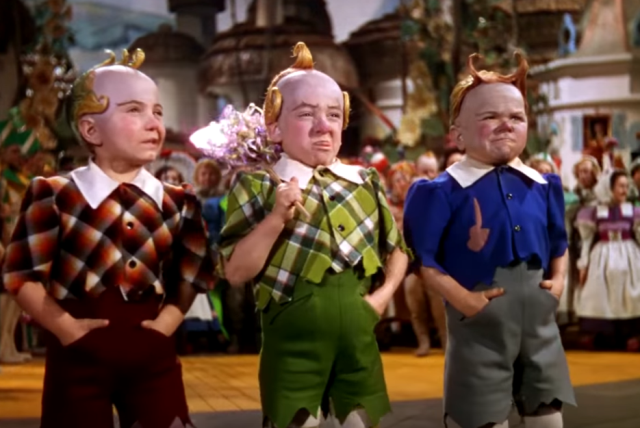 Still from the
1939 MGM film “The Wizard of Oz”.  Three midgets, dressed respectively
in bright red, green, and blue suits, look simultaneously like
overgrown babies and like weirdly shrunken old men.  They are bald
except for fancifully curled golden mohawks that resemble gilded
staircase bannisters, and are making contorted faces.