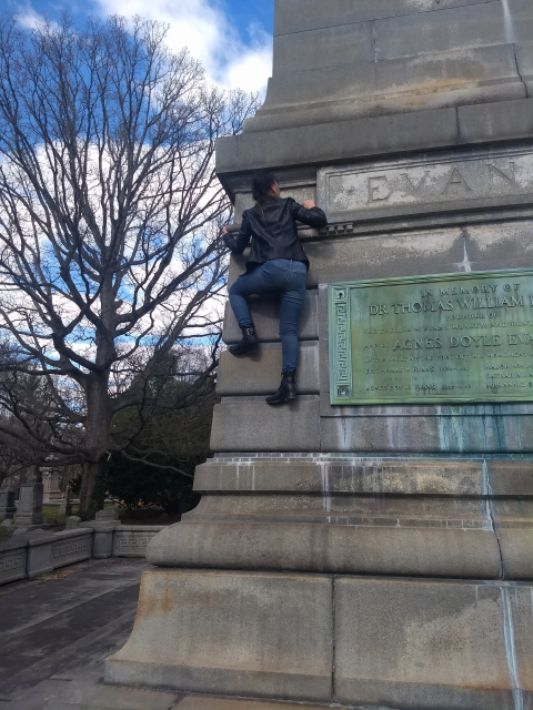 A young girl, seen from the back, is climbing a large stone
monument.  She is wearing black boots, blue jeans, and a black leather
jacket.  She is about six feet off the ground. Attached to the monument to her right is a
green copper plate that says  (among other things) ‘In memory of
DR. THOMAS WILLIAM EVANS’.  In the background is a tree, and other, smaller
monuments can be seen.
