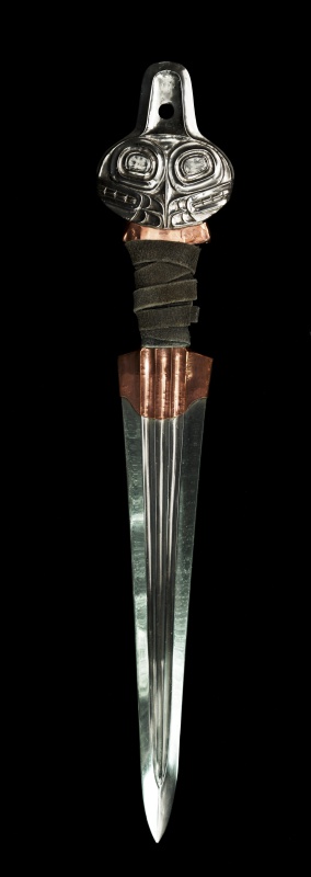 The Killer Whale dagger against
a black background.  It is shiny steel with copper overlay and leather
wrapping about the grip area. The blade is a long, tapered triangular
form with three prominent flutes down the center of its length. The
integral steel pommel is relief-formed into the image of two orca
whale heads looking outward with a single dorsal fin extending upward
from the whale heads. A single cut hole pierces the dorsal fin. The
pommel is flat on the reverse side