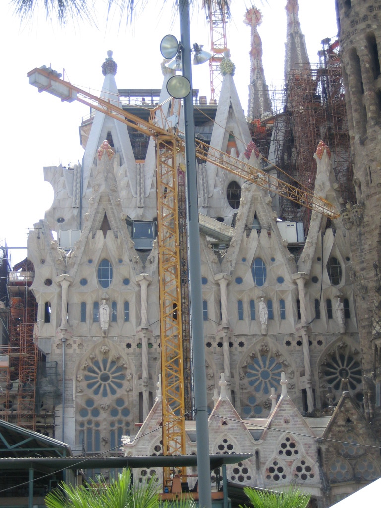 Outside of Sagrada Família, clearly under
construction: part of the structure is covered with scaffolding, and
there is a big yellow construction crane right in the middle of the
picture.  Behind this the wall of the building is divided into
sections, each with a tall stone window divided into circles and
flower shapes; above that a sculpture of a saint, above that an oval
window, an then a steeply pointed stone roof.  Each steep point is
capped by a four-armed basket of colored stone globes resembling
fruits of various colors: the leftmost ones are warm oranges and
yellows; to the right are smaller but more numerous reds and purples.
Behind this is another similar row of roodsm even higher, with more
fruits like enormous bunches of purple and green grapes.