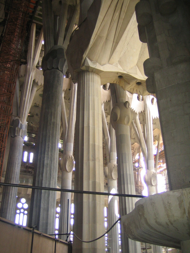 Interior of Sagrada Família, still under
construction: walls and parts of the roof are missing, windows lack
stained glass, scaffolding is visible at left.  The roof is held up by
fluted stone columns the resemble tree trunks.  Far overhead the tree
trunks are decorated with huge stone boles and knots, and above the boles the
columns divide into branches on which hang stone foliage.