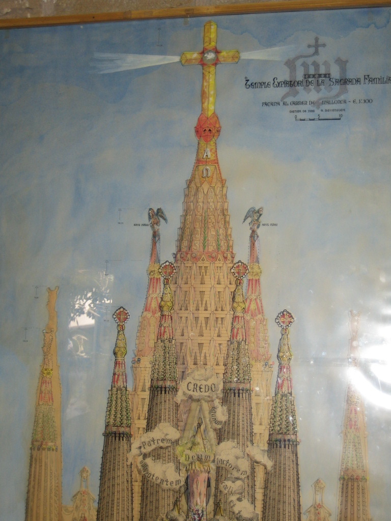 A colored painting of one side
of the building, as it was imagined around 1902.  At the bottom, we
see that main entrance is decorated with clouds labeled “CREDO”,
“Patrem”, “Deum”, and so on.  Four of the apostles” towers are around
this.  Behind these we can see two of the evangelists’ towers,
surmounted by a winged lion (for Mark) and an eagle (for John).
Behind these, in the center, and by far the largest, is the conical
tower of Jesus, surmounted by an immense golden cross, from whose arms
stream rays of light.  Under this the pointed top of the tower carries
the Greek letters alpha and omega, and under this are columns of stone
panels with more letters on them.