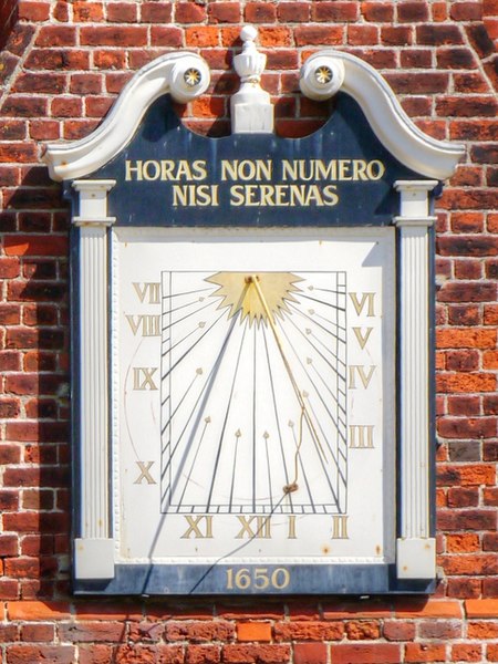 wooden sundial mounted on a brick wall; the numbers run counter-clockwise down the left edge from VII to X, then along the bottom edge from XI through XII to II, and then up the right edge from III to VI.