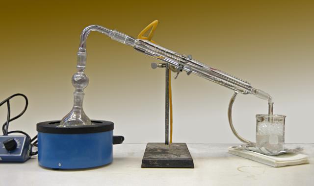 A chemistry lab
apparatus.  In the middle thje the Liebig condenser, a pair of long
concentric glass tubes, one inside the other, supported by a metal
ring stand.  At left a heater heats a flash whose top is connected to
the upper end of the inner tube of the condenser.  The condensate
collects in a flask at right.  Two rubber tubes connect to the top and
bottom of the outer tube, carrying water through it.