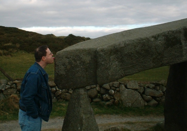 A slightly balding dark-haired man with glasses is leaning slightly as he sticks out his tongue to touch a massive rectangular stone that is resting at head height atop smaller upright stones.  In the background are a green hill and a stone wall.  The man has his hands in the pockets of his blue jeans and is wearing a blue denom jacket.