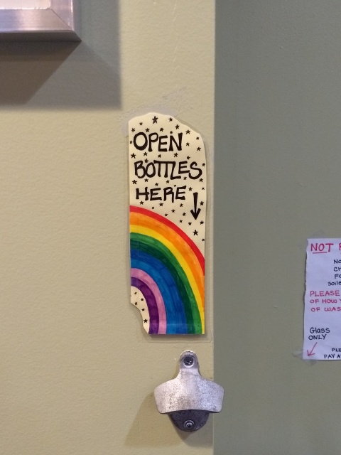 A sign on a wall-mounted
bottle opener with a picture of a rainbow and the words “OPEN BOTTLES HERE”