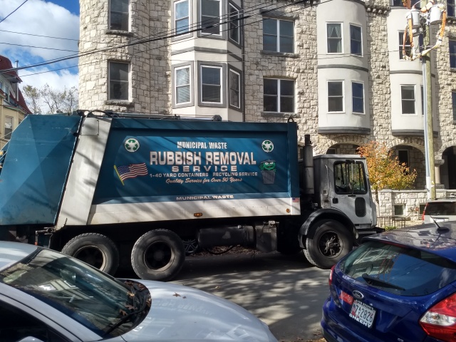 The side of a blue trash
truck, proclaiming “MUNICIPAL WASTE”, “RUBBISH REMOVAL”, and
“RECYCLING SERVICE”.  The truck is also emblazoned with three-arrow
recycling symbols, a waving U.S. flag, and a portrait of a
cheerful-looking Oscar the Grouch emerging from his trash can.