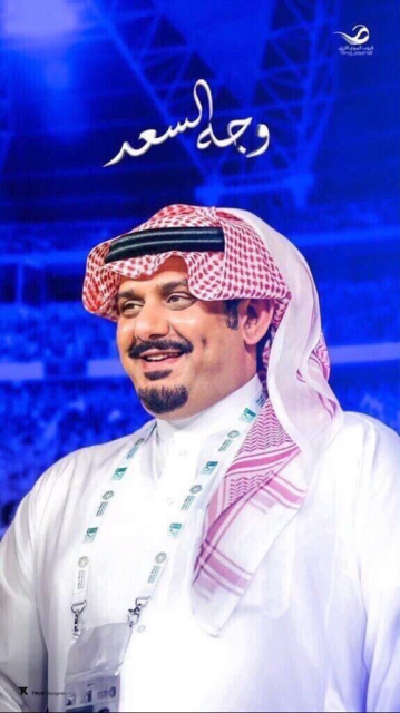 A smiling, shiny-cheeked Arabian
man in middle age, with a black mustache and goatee.  He wears a very
typical red-and-white checked head scarf (ghutra), held in place by a
black double cord (igal).  The background is an indistinct view of a
crowd in a sport stadium, in monochrome royal blue.